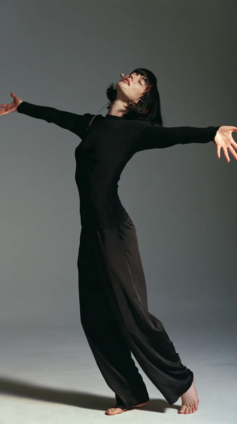 full-length shot, a Caucasian American woman in her late twenties with pale skin and a glossy black blunt bob with heavy straight bangs and one clay-orange streak, a thin headset microphone, in a black ribbed…