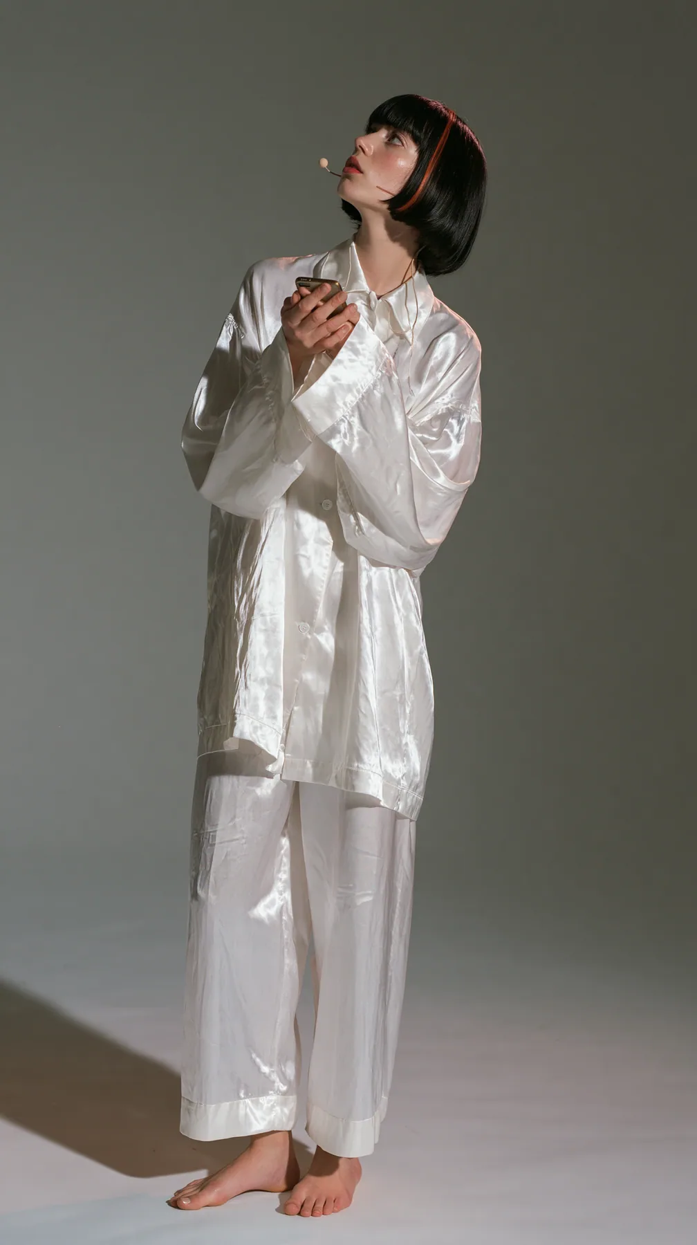 full-length shot, a Caucasian American woman in her late twenties with pale skin and a glossy black blunt bob with heavy straight bangs and one clay-orange streak, a thin headset microphone, in oversized white…