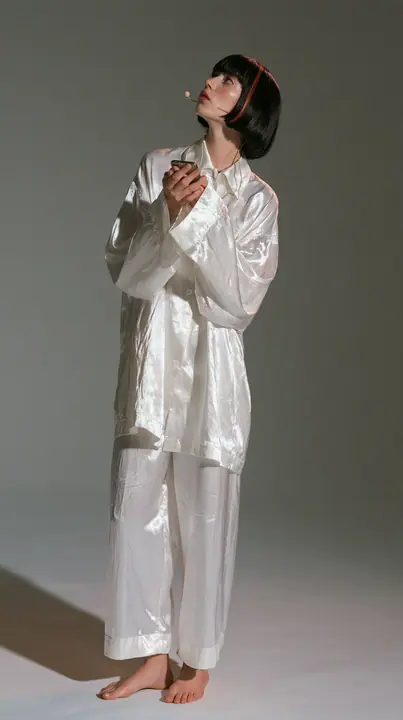 full-length shot, a Caucasian American woman in her late twenties with pale skin and a glossy black blunt bob with heavy straight bangs and one clay-orange streak, a thin headset microphone, in oversized white…