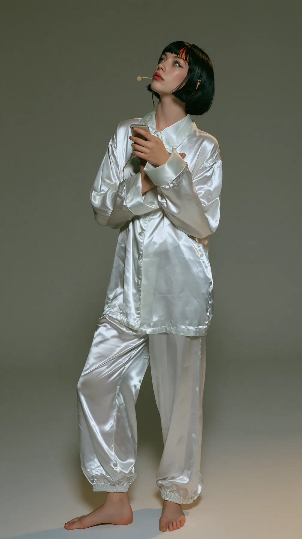 full-length shot, a Caucasian American woman in her late twenties with pale skin and a glossy black blunt bob with heavy straight bangs and one clay-orange streak, a thin headset microphone, in oversized white…