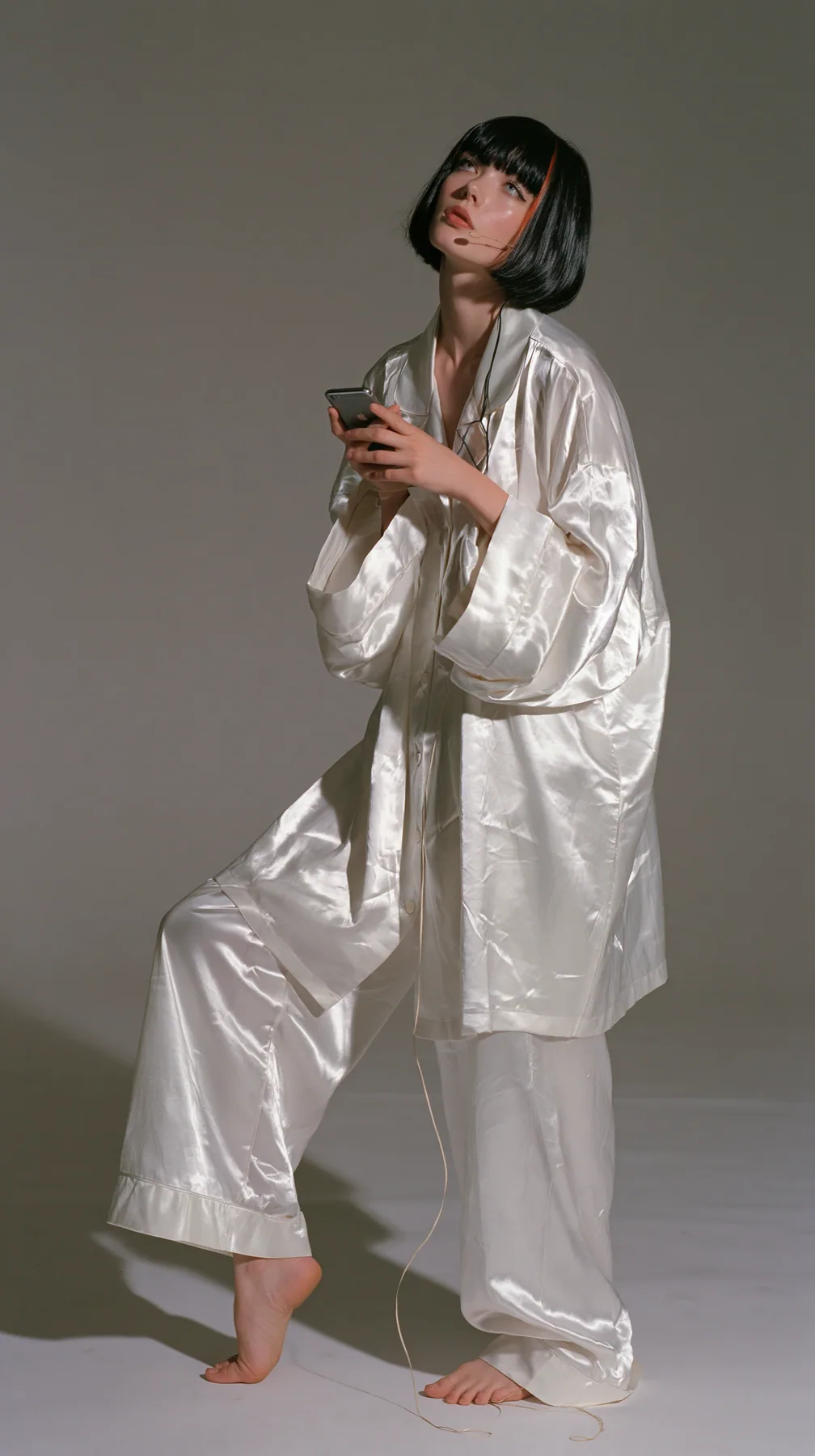 full-length shot, a Caucasian American woman in her late twenties with pale skin and a glossy black blunt bob with heavy straight bangs and one clay-orange streak, a thin headset microphone, in oversized white…