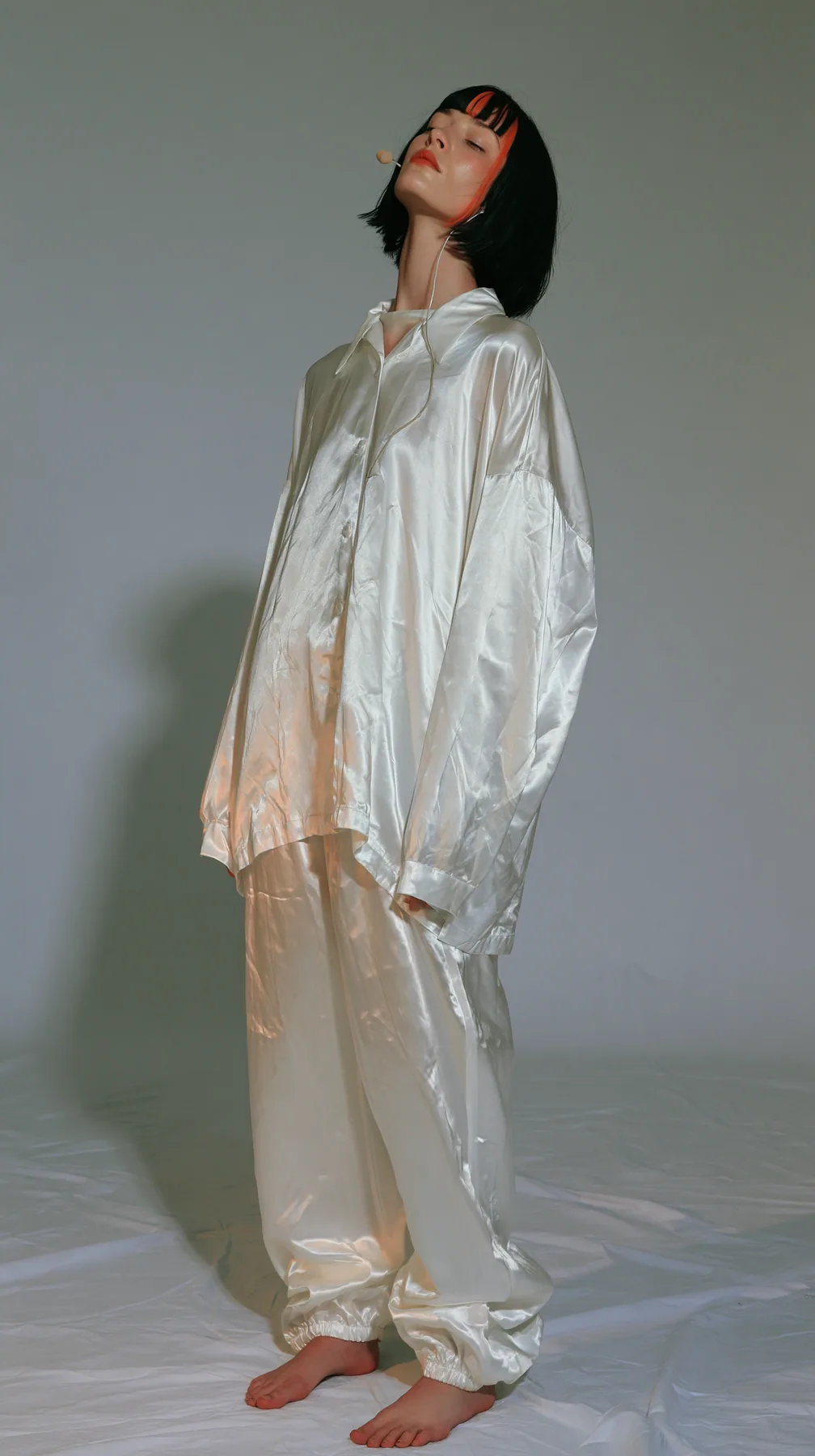 full-length shot, a Caucasian American woman in her late twenties with pale skin and a glossy black blunt bob with heavy straight bangs and one clay-orange streak, a thin headset microphone, in oversized white…