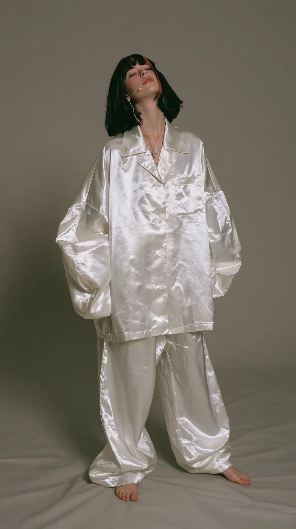 full-length shot, a Caucasian American woman in her late twenties with pale skin and a glossy black blunt bob with heavy straight bangs and one clay-orange streak, a thin headset microphone, in oversized white…