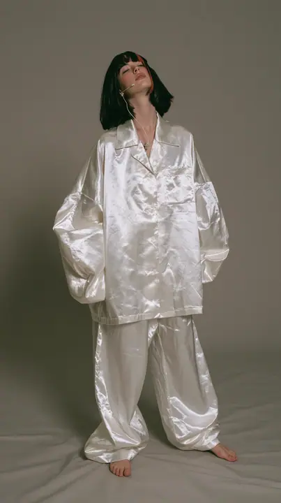 full-length shot, a Caucasian American woman in her late twenties with pale skin and a glossy black blunt bob with heavy straight bangs and one clay-orange streak, a thin headset microphone, in oversized white…
