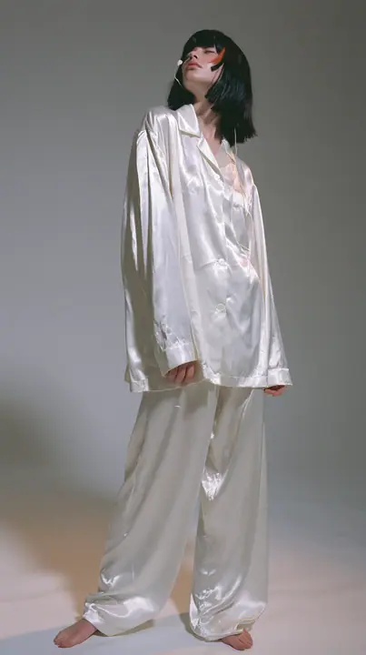 full-length shot, a Caucasian American woman in her late twenties with pale skin and a glossy black blunt bob with heavy straight bangs and one clay-orange streak, a thin headset microphone, in oversized white…
