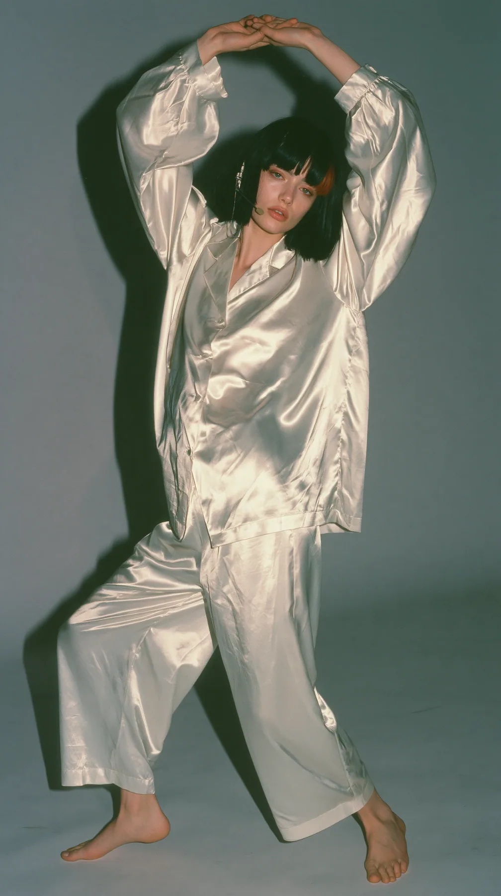 full-length shot, a Caucasian American woman in her late twenties with pale skin and a glossy black blunt bob with heavy straight bangs and one clay-orange streak, a thin headset microphone, in oversized white…
