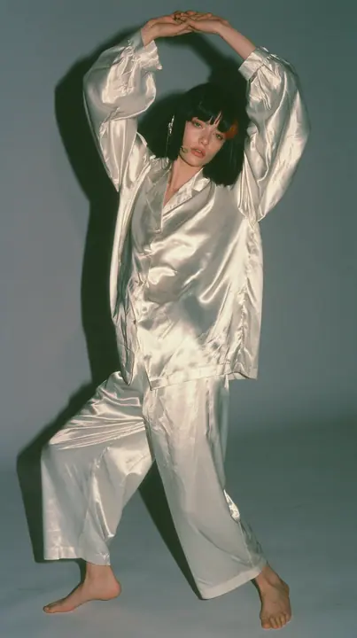 full-length shot, a Caucasian American woman in her late twenties with pale skin and a glossy black blunt bob with heavy straight bangs and one clay-orange streak, a thin headset microphone, in oversized white…