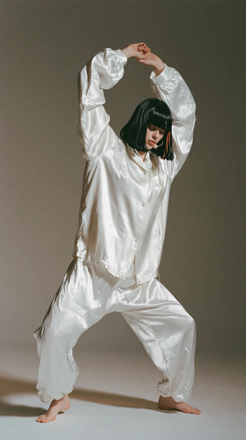 full-length shot, a Caucasian American woman in her late twenties with pale skin and a glossy black blunt bob with heavy straight bangs and one clay-orange streak, a thin headset microphone, in oversized white…