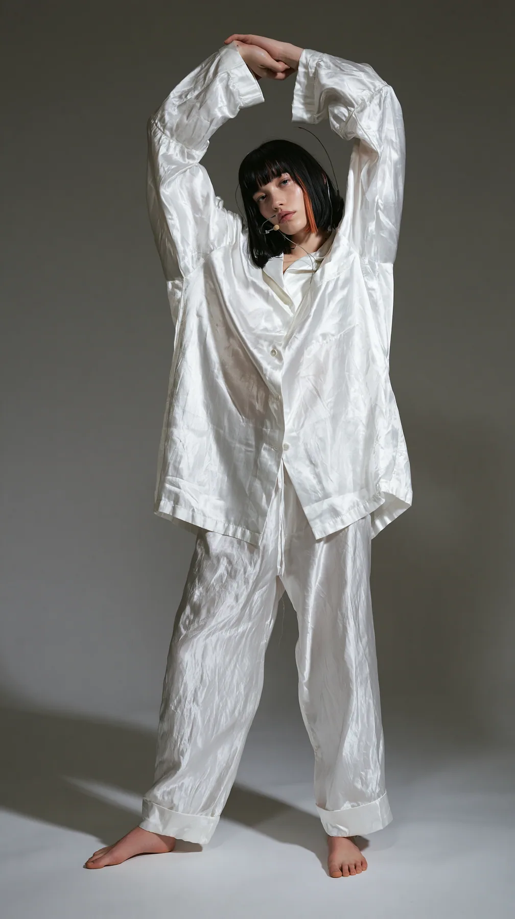 full-length shot, a Caucasian American woman in her late twenties with pale skin and a glossy black blunt bob with heavy straight bangs and one clay-orange streak, a thin headset microphone, in oversized white…