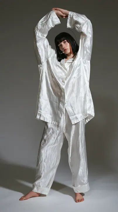 full-length shot, a Caucasian American woman in her late twenties with pale skin and a glossy black blunt bob with heavy straight bangs and one clay-orange streak, a thin headset microphone, in oversized white…