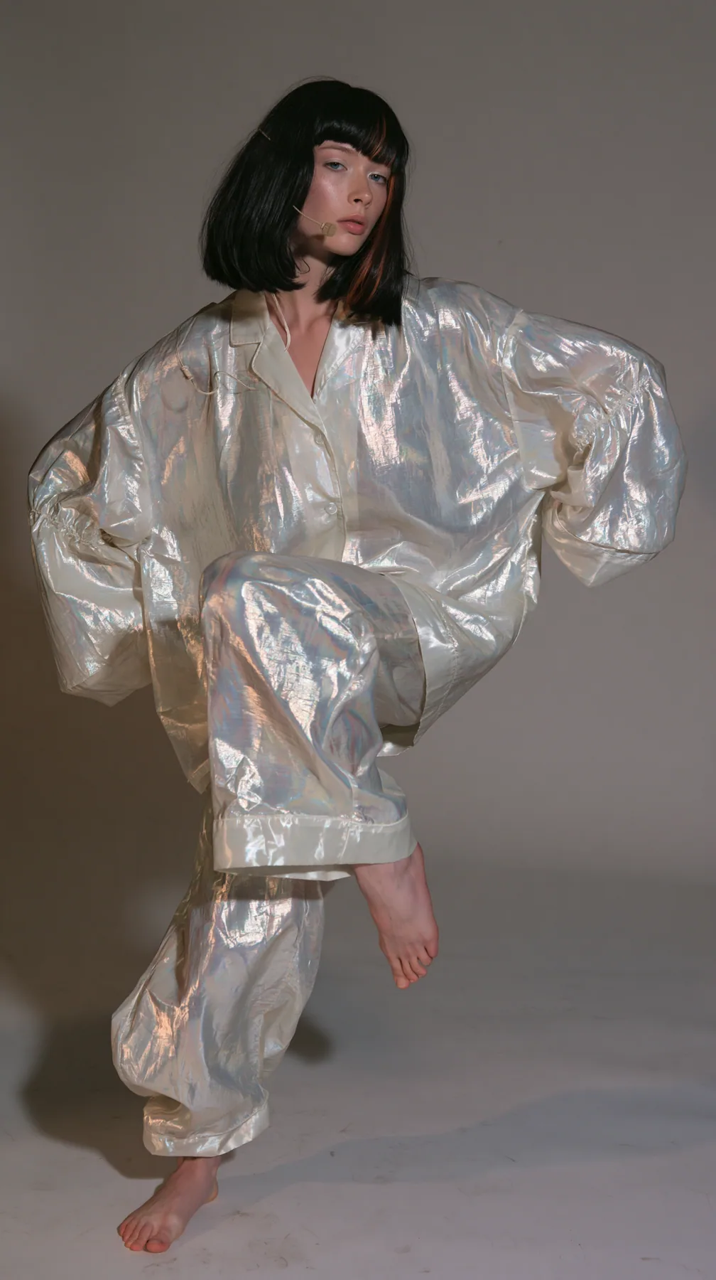 full-length shot, a Caucasian American woman in her late twenties with pale skin and a glossy black blunt bob with heavy straight bangs and one clay-orange streak, a thin headset microphone, in oversized iridescent…