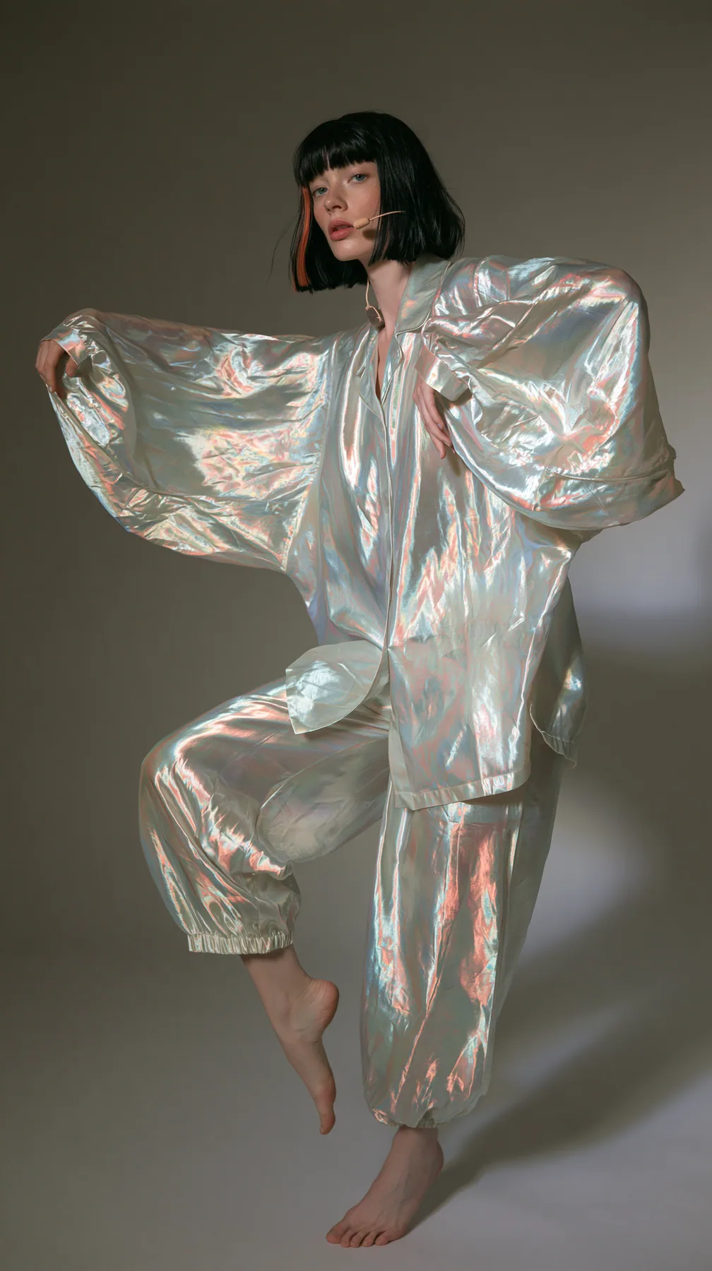 full-length shot, a Caucasian American woman in her late twenties with pale skin and a glossy black blunt bob with heavy straight bangs and one clay-orange streak, a thin headset microphone, in oversized iridescent…