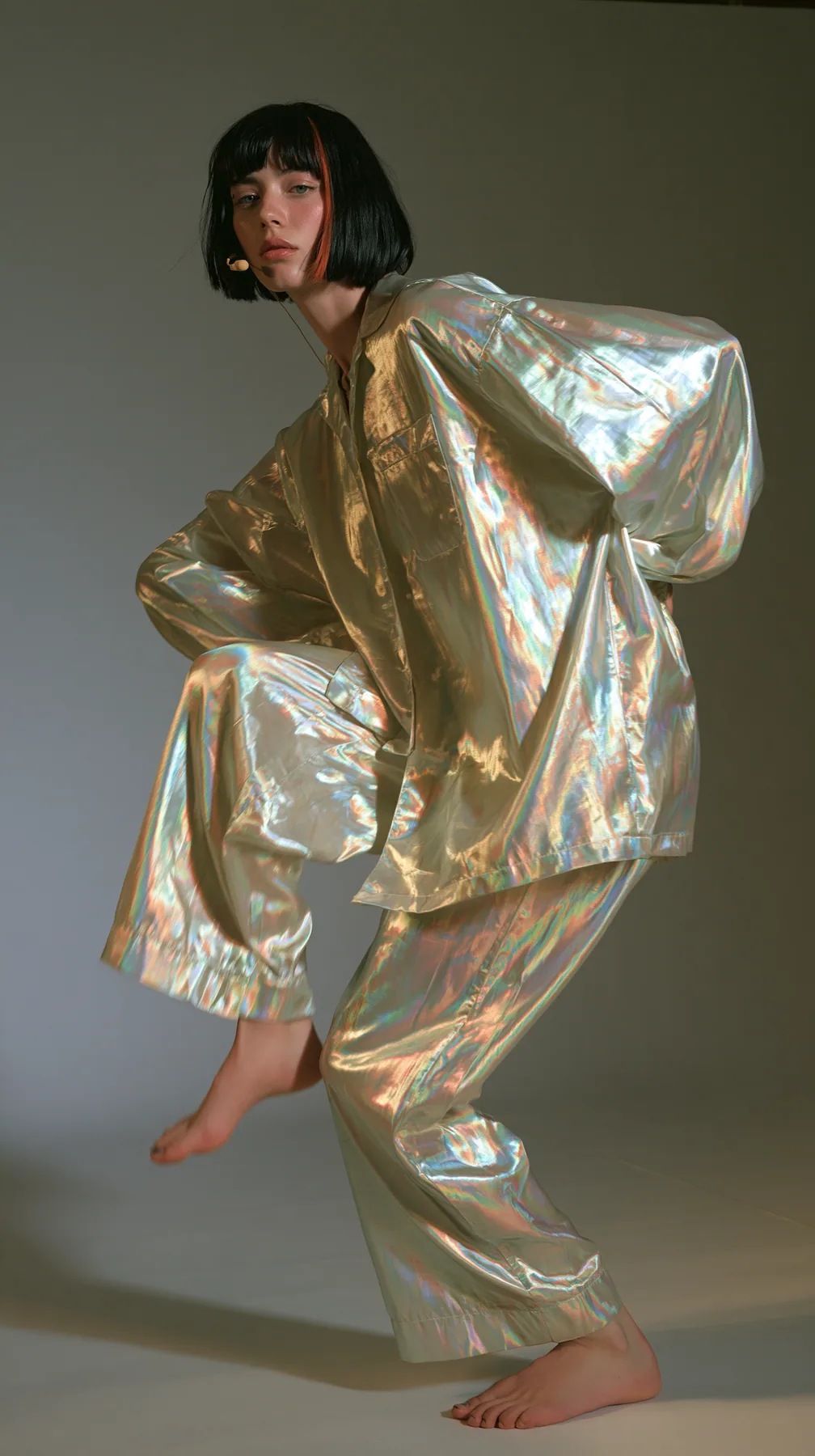 full-length shot, a Caucasian American woman in her late twenties with pale skin and a glossy black blunt bob with heavy straight bangs and one clay-orange streak, a thin headset microphone, in oversized iridescent…