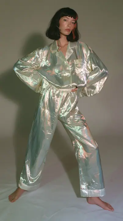 full-length shot, a Caucasian American woman in her late twenties with pale skin and a glossy black blunt bob with heavy straight bangs and one clay-orange streak, a thin headset microphone, in oversized iridescent…
