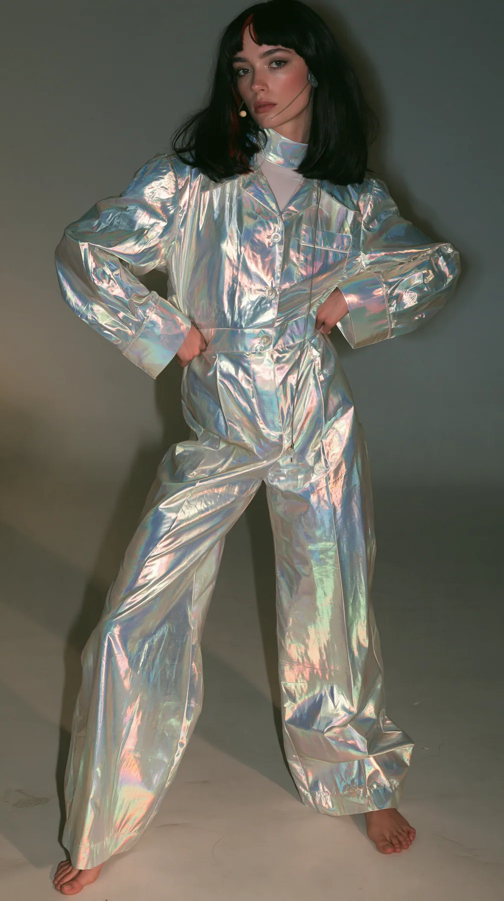 full-length shot, a Caucasian American woman in her late twenties with pale skin and a glossy black blunt bob with heavy straight bangs and one clay-orange streak, a thin headset microphone, in oversized iridescent…