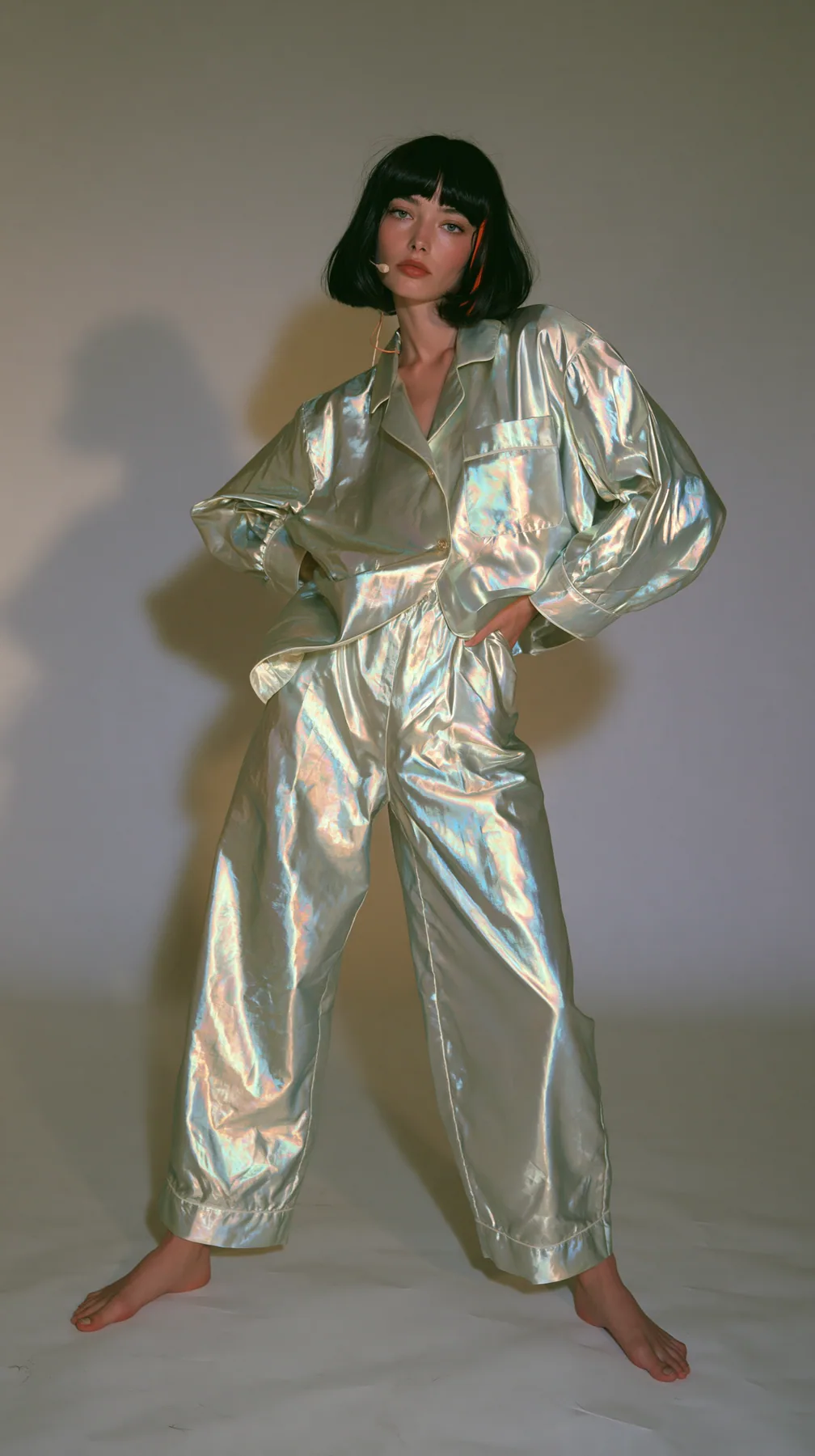 full-length shot, a Caucasian American woman in her late twenties with pale skin and a glossy black blunt bob with heavy straight bangs and one clay-orange streak, a thin headset microphone, in oversized iridescent…