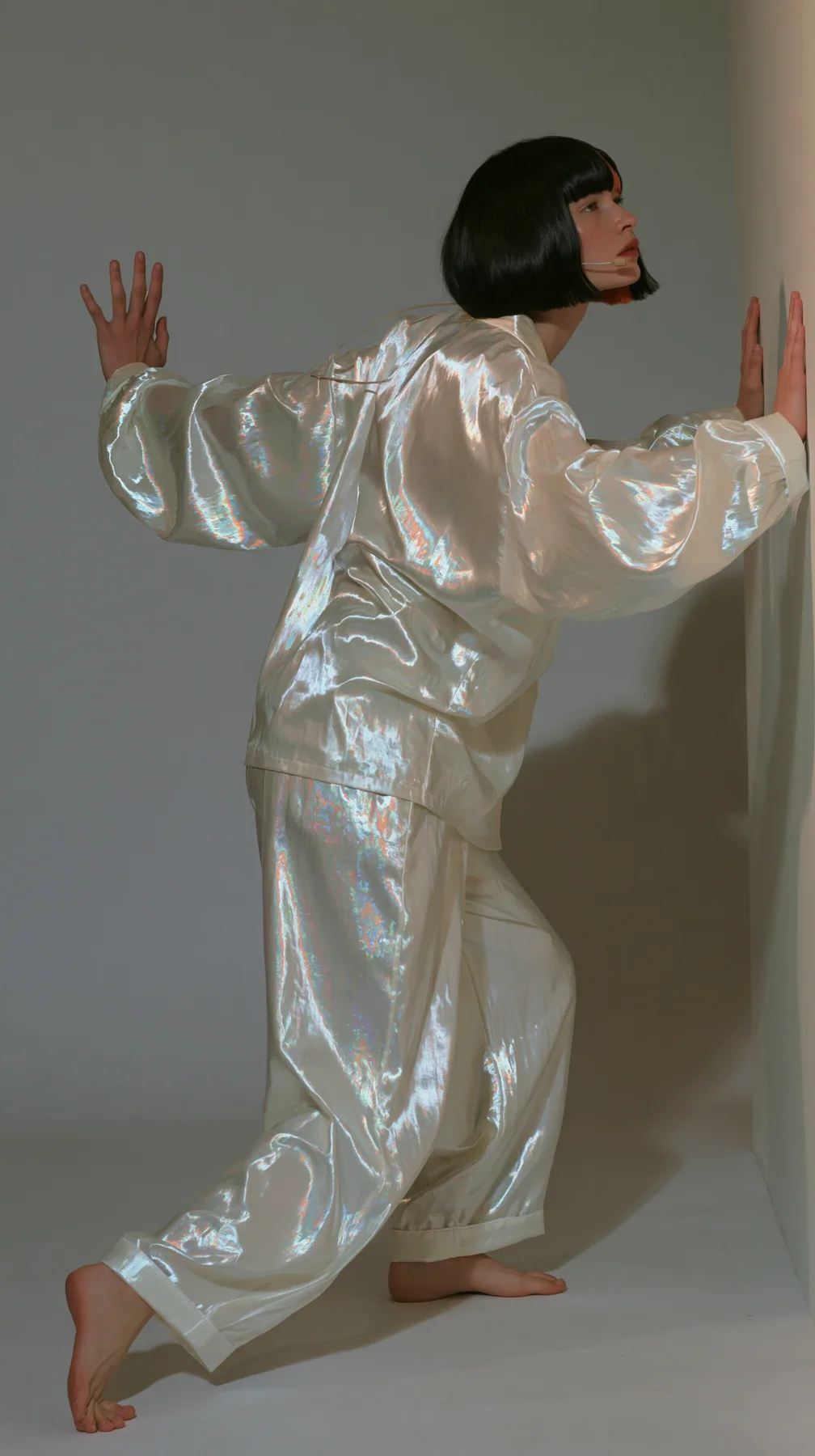 full-length side view, a Caucasian American woman in her late twenties with pale skin and a glossy black blunt bob with heavy straight bangs and one clay-orange streak, a thin headset microphone, in oversized…