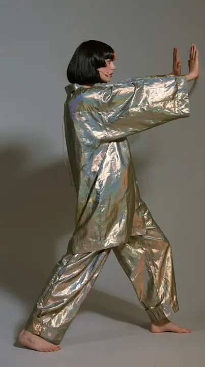 full-length side view, a Caucasian American woman in her late twenties with pale skin and a glossy black blunt bob with heavy straight bangs and one clay-orange streak, a thin headset microphone, in oversized…