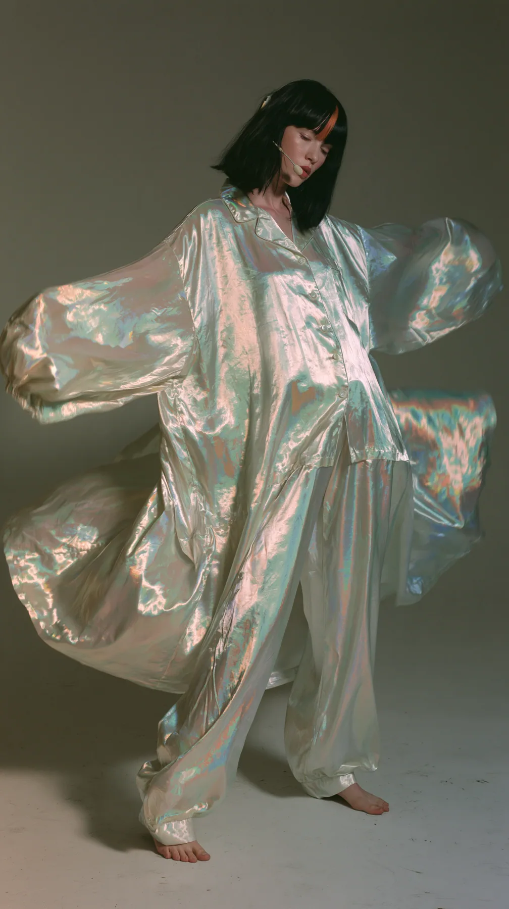 full-length shot, a Caucasian American woman in her late twenties with pale skin and a glossy black blunt bob with heavy straight bangs and one clay-orange streak, a thin headset microphone, in oversized iridescent…