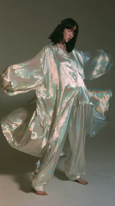 full-length shot, a Caucasian American woman in her late twenties with pale skin and a glossy black blunt bob with heavy straight bangs and one clay-orange streak, a thin headset microphone, in oversized iridescent…
