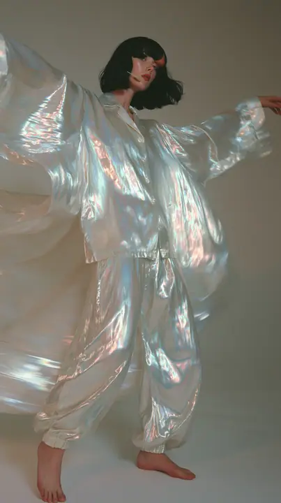full-length shot, a Caucasian American woman in her late twenties with pale skin and a glossy black blunt bob with heavy straight bangs and one clay-orange streak, a thin headset microphone, in oversized iridescent…