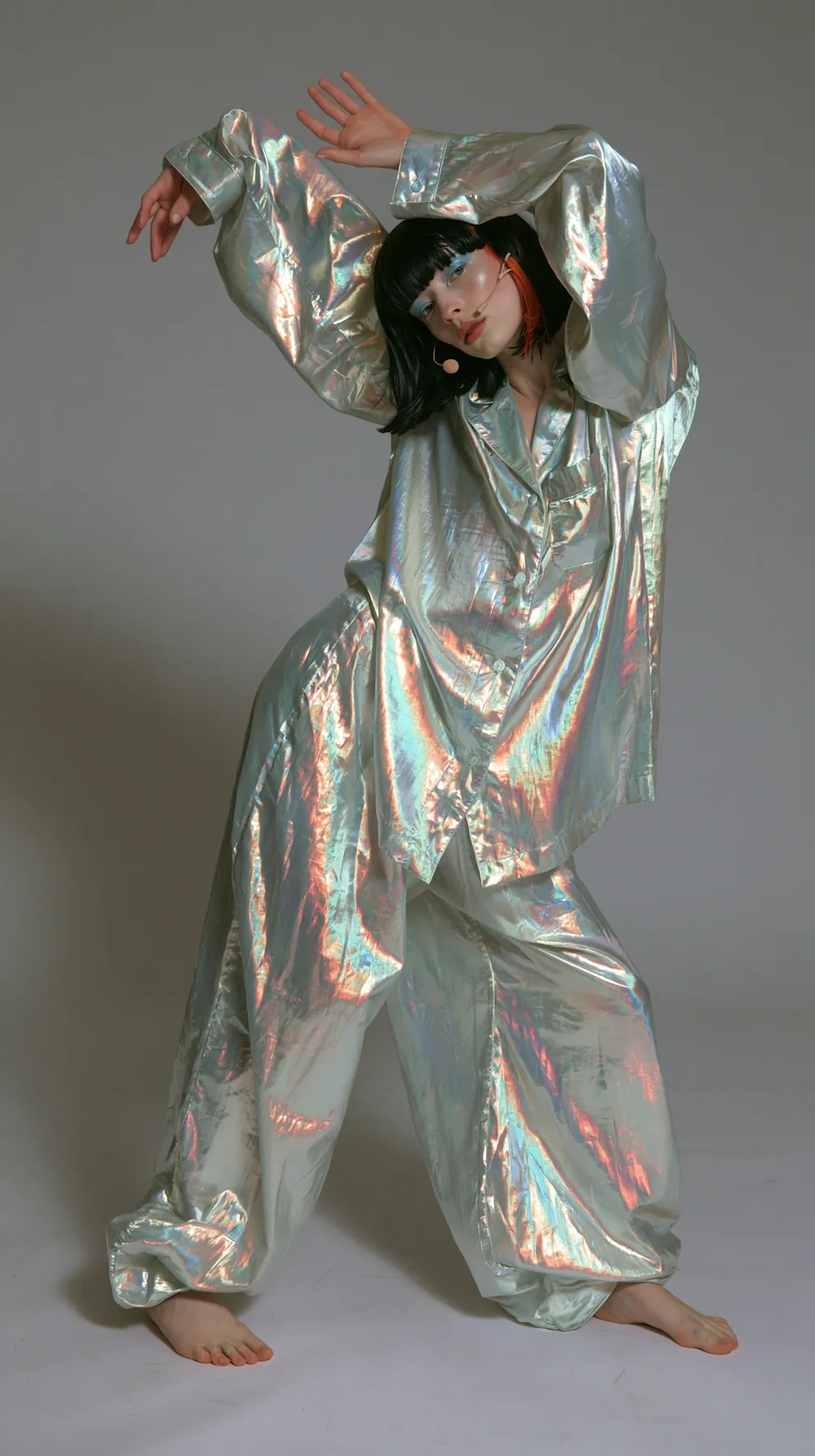 full-length shot, a Caucasian American woman in her late twenties with pale skin and a glossy black blunt bob with heavy straight bangs and one clay-orange streak, a thin headset microphone, in oversized iridescent…
