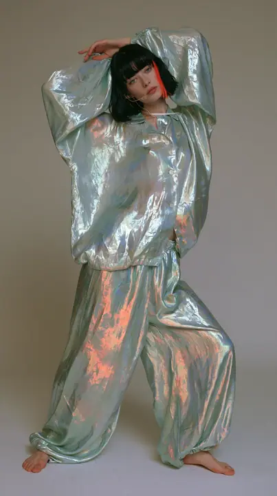 full-length shot, a Caucasian American woman in her late twenties with pale skin and a glossy black blunt bob with heavy straight bangs and one clay-orange streak, a thin headset microphone, in oversized iridescent…