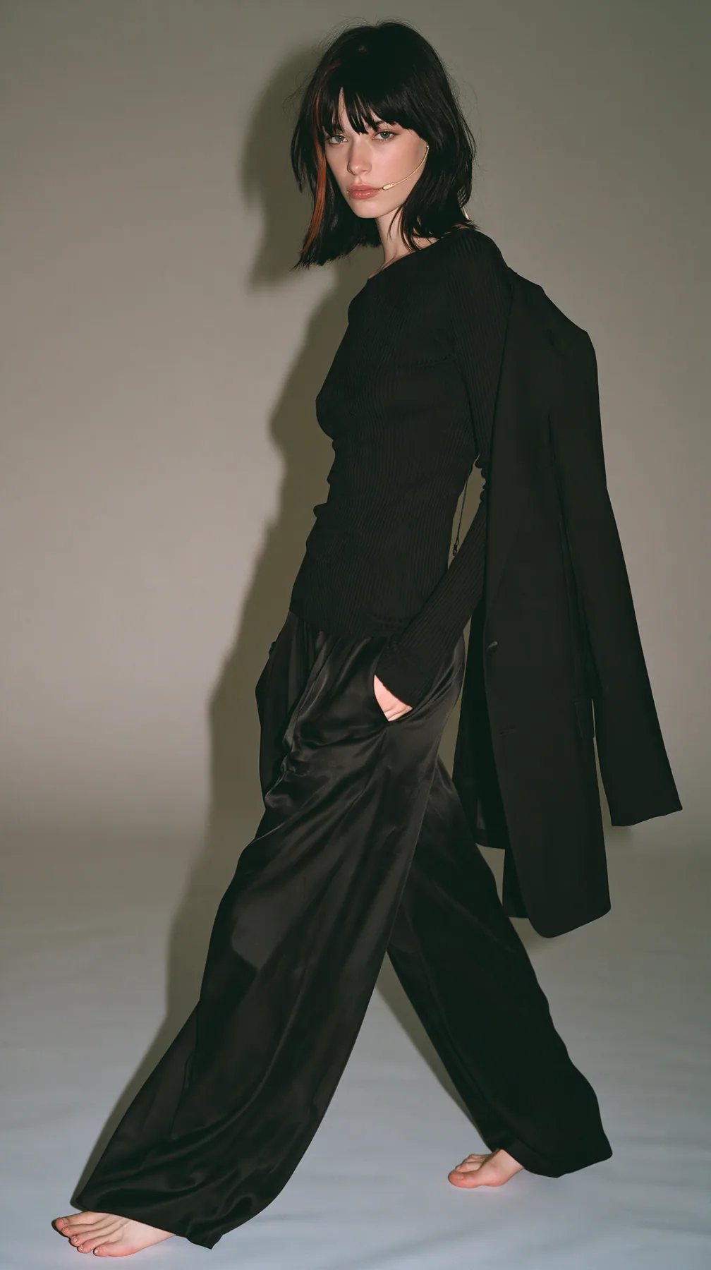 full-length shot, a Caucasian American woman in her late twenties with pale skin and a glossy black blunt bob with heavy straight bangs and one clay-orange streak, a thin headset microphone, in a black ribbed…