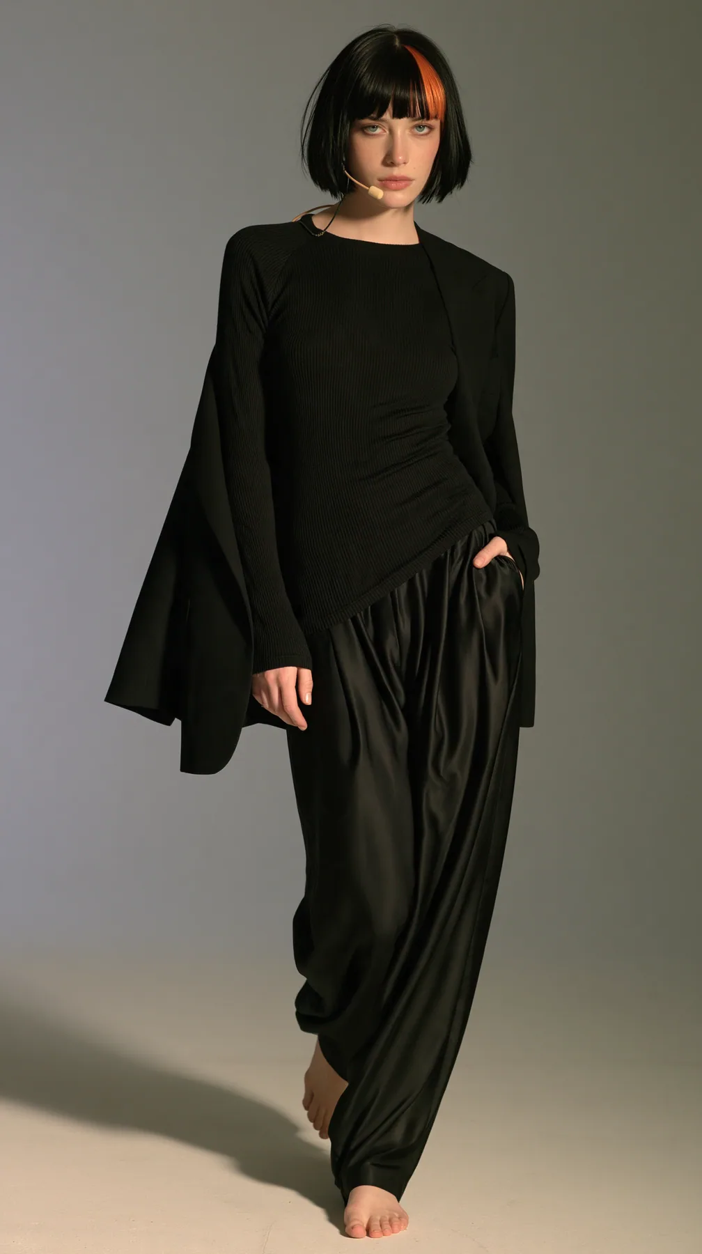 full-length shot, a Caucasian American woman in her late twenties with pale skin and a glossy black blunt bob with heavy straight bangs and one clay-orange streak, a thin headset microphone, in a black ribbed…