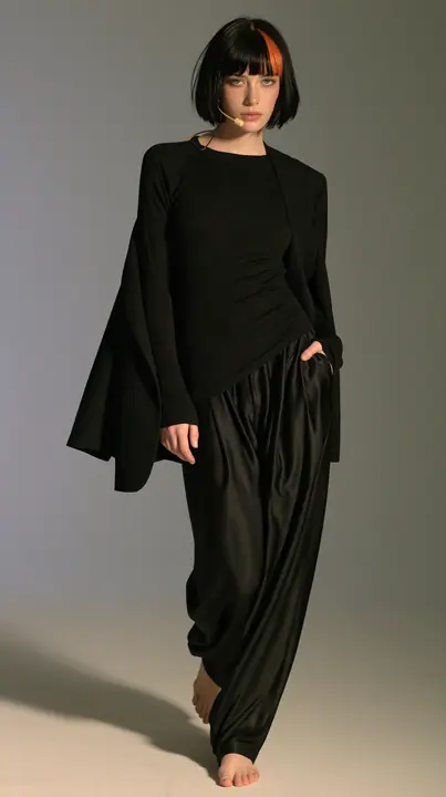 full-length shot, a Caucasian American woman in her late twenties with pale skin and a glossy black blunt bob with heavy straight bangs and one clay-orange streak, a thin headset microphone, in a black ribbed…