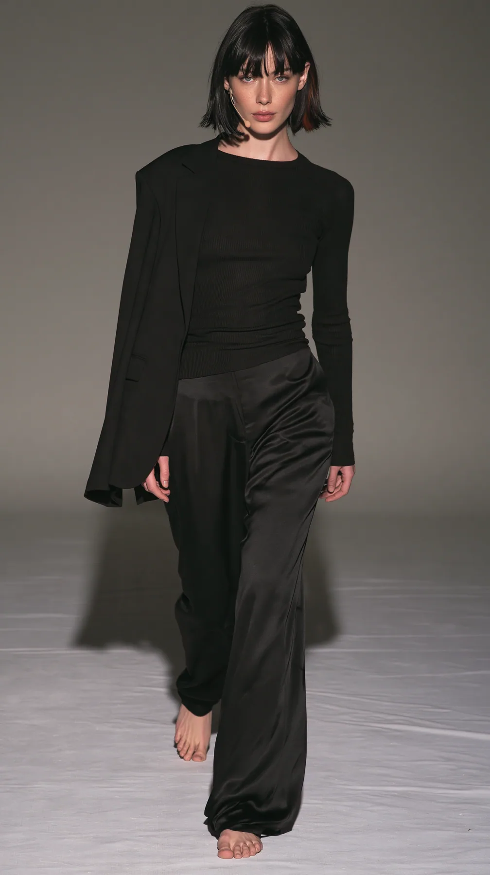 full-length shot, a Caucasian American woman in her late twenties with pale skin and a glossy black blunt bob with heavy straight bangs and one clay-orange streak, a thin headset microphone, in a black ribbed…