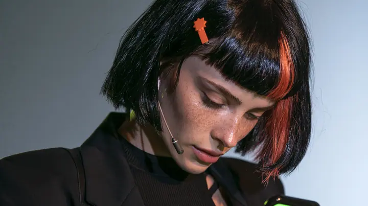 over-the-shoulder medium close-up, a 28-year-old Caucasian American woman with a grown-up angular face, defined cheekbones and a strong jawline, pale skin and light freckles, a glossy black blunt jaw-length bob with…