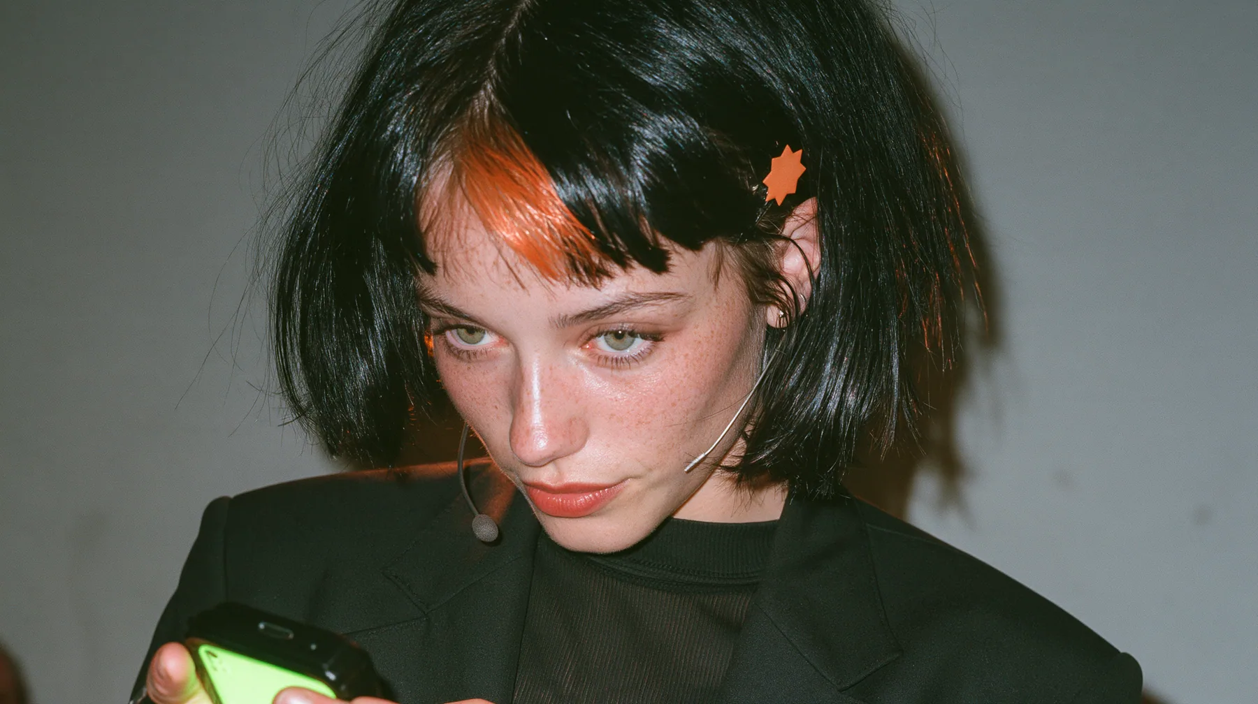 over-the-shoulder medium close-up, a 28-year-old Caucasian American woman with a grown-up angular face, defined cheekbones and a strong jawline, pale skin and light freckles, a glossy black blunt jaw-length bob with…