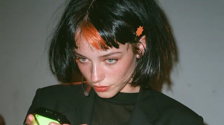 over-the-shoulder medium close-up, a 28-year-old Caucasian American woman with a grown-up angular face, defined cheekbones and a strong jawline, pale skin and light freckles, a glossy black blunt jaw-length bob with…