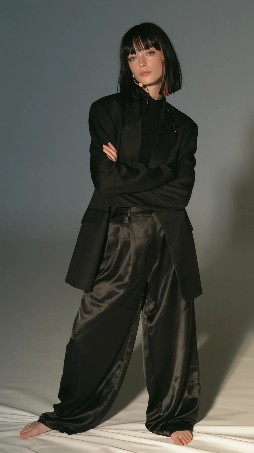 full-length shot, a Caucasian American woman in her late twenties with pale skin and a glossy black blunt bob with heavy straight bangs and one clay-orange streak, a thin headset microphone, in a sharp black blazer…