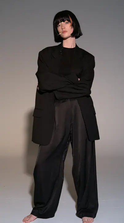 full-length shot, a Caucasian American woman in her late twenties with pale skin and a glossy black blunt bob with heavy straight bangs and one clay-orange streak, a thin headset microphone, in a sharp black blazer…