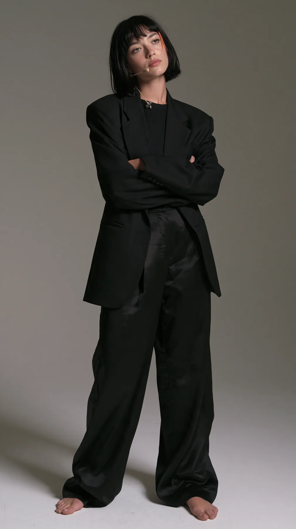 full-length shot, a Caucasian American woman in her late twenties with pale skin and a glossy black blunt bob with heavy straight bangs and one clay-orange streak, a thin headset microphone, in a sharp black blazer…