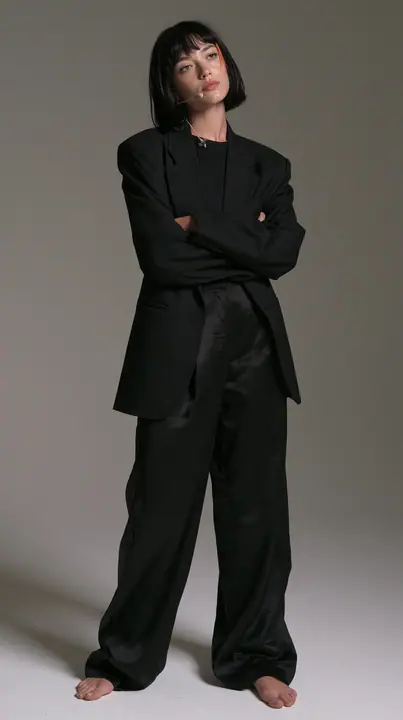 full-length shot, a Caucasian American woman in her late twenties with pale skin and a glossy black blunt bob with heavy straight bangs and one clay-orange streak, a thin headset microphone, in a sharp black blazer…