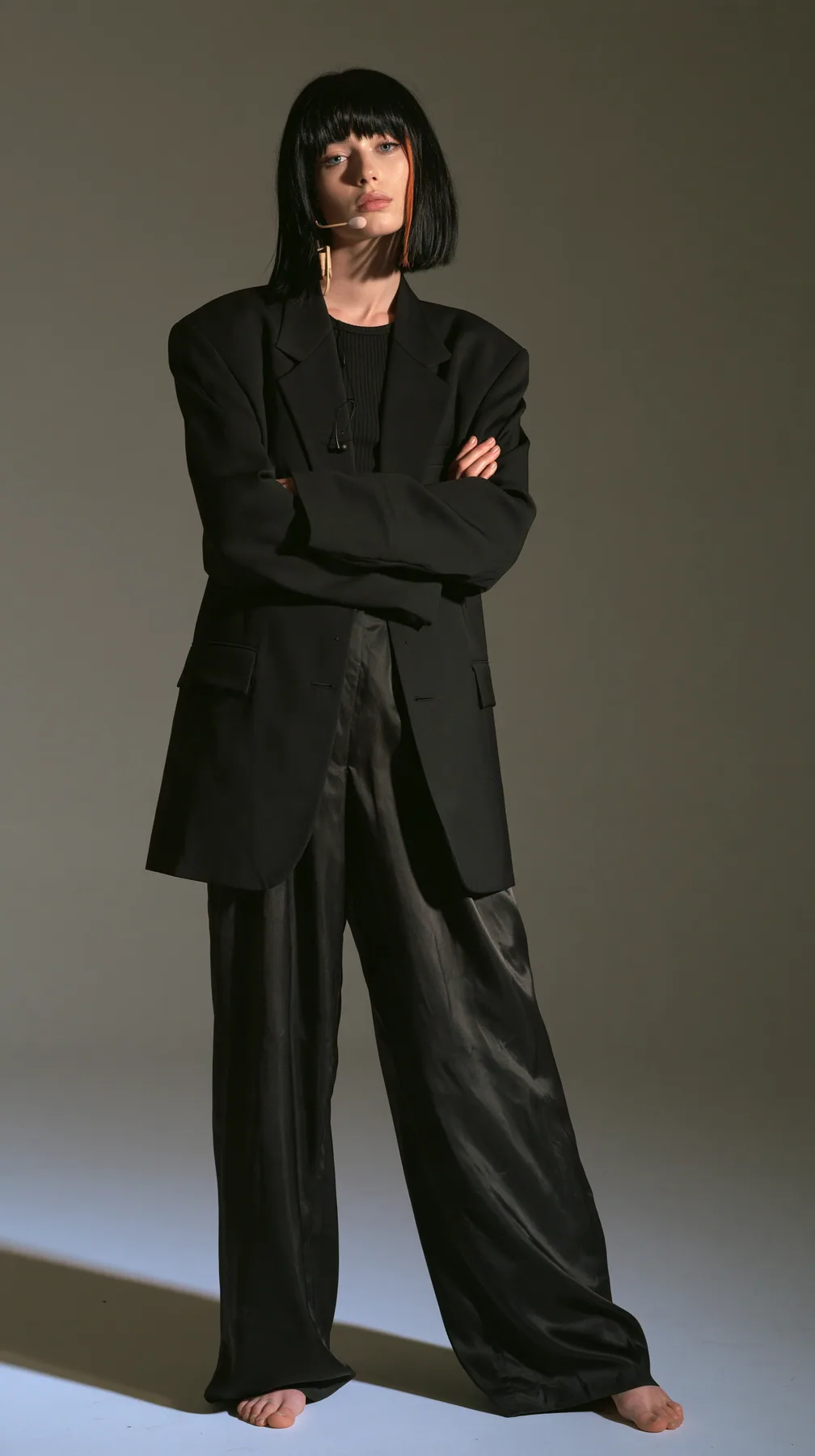 full-length shot, a Caucasian American woman in her late twenties with pale skin and a glossy black blunt bob with heavy straight bangs and one clay-orange streak, a thin headset microphone, in a sharp black blazer…