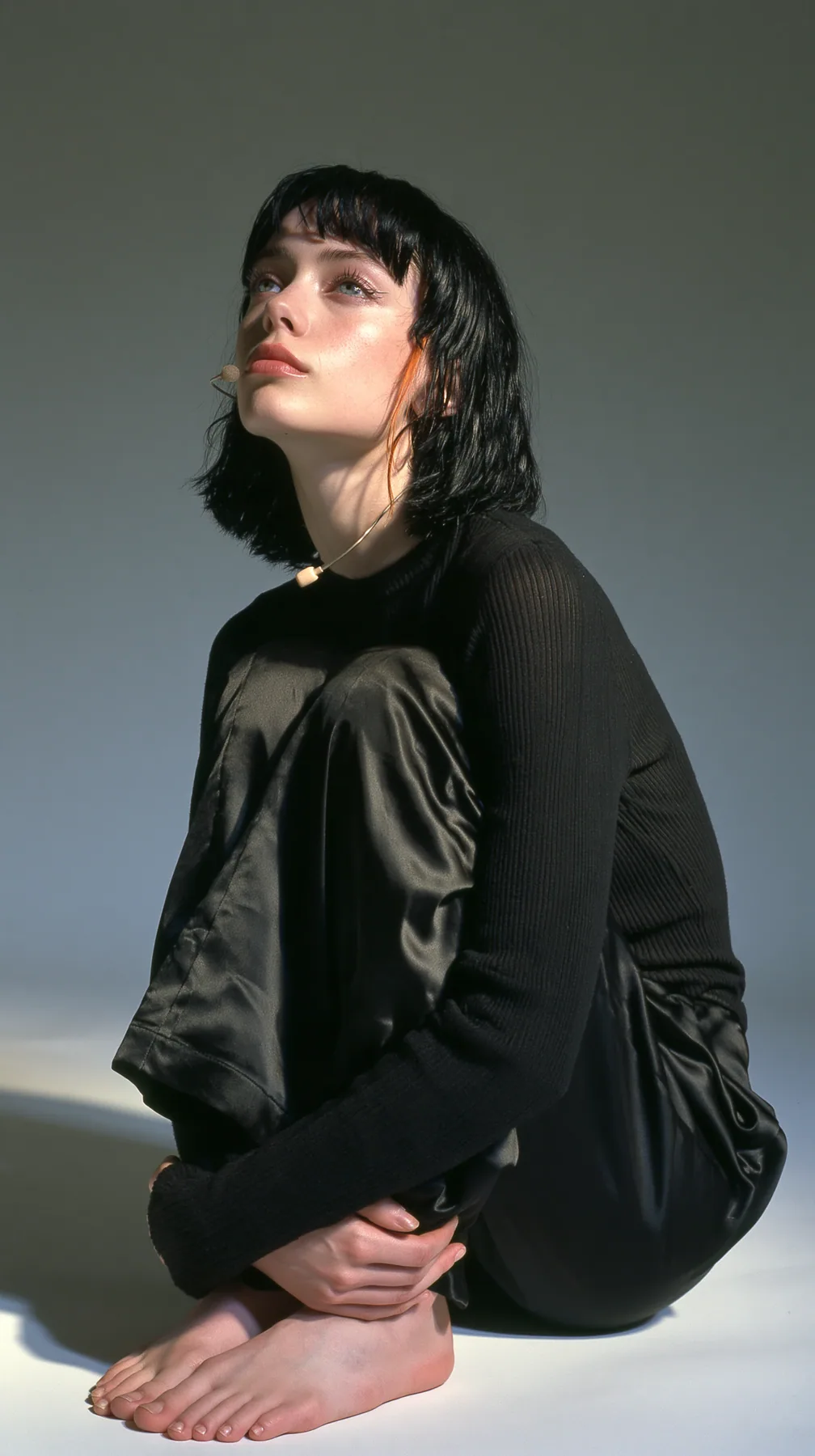 full-length shot, a Caucasian American woman in her late twenties with pale skin and a glossy black blunt bob with heavy straight bangs and one clay-orange streak, a thin headset microphone, in a black ribbed…