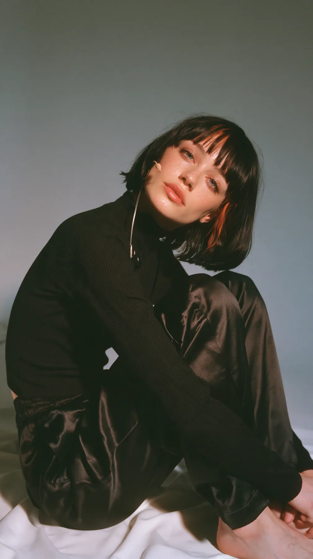full-length shot, a Caucasian American woman in her late twenties with pale skin and a glossy black blunt bob with heavy straight bangs and one clay-orange streak, a thin headset microphone, in a black ribbed…