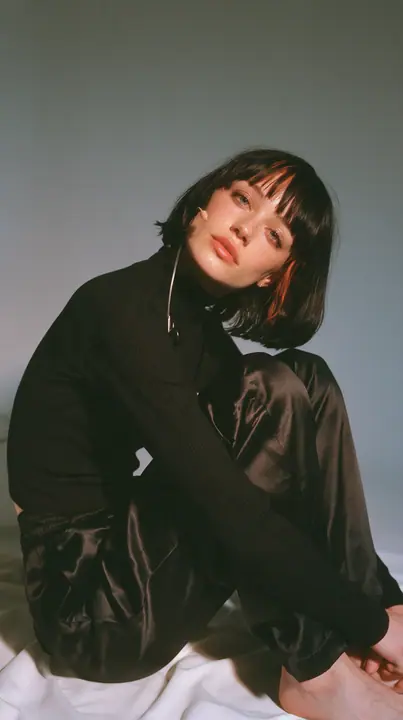 full-length shot, a Caucasian American woman in her late twenties with pale skin and a glossy black blunt bob with heavy straight bangs and one clay-orange streak, a thin headset microphone, in a black ribbed…