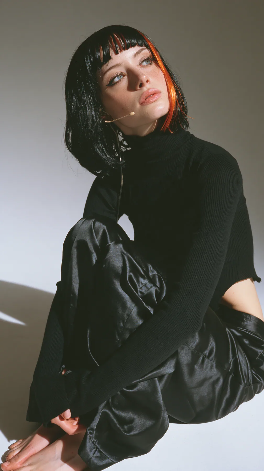 full-length shot, a Caucasian American woman in her late twenties with pale skin and a glossy black blunt bob with heavy straight bangs and one clay-orange streak, a thin headset microphone, in a black ribbed…