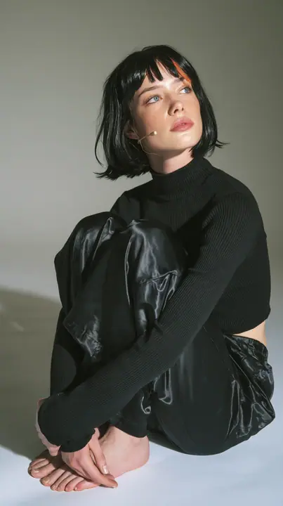full-length shot, a Caucasian American woman in her late twenties with pale skin and a glossy black blunt bob with heavy straight bangs and one clay-orange streak, a thin headset microphone, in a black ribbed…
