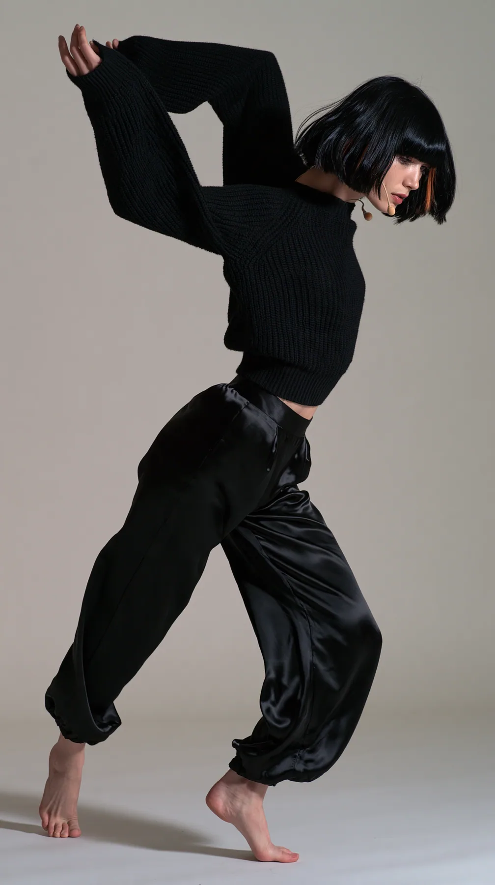 full-length shot, a Caucasian American woman in her late twenties with pale skin and a glossy black blunt bob with heavy straight bangs and one clay-orange streak, a thin headset microphone, in a black ribbed…
