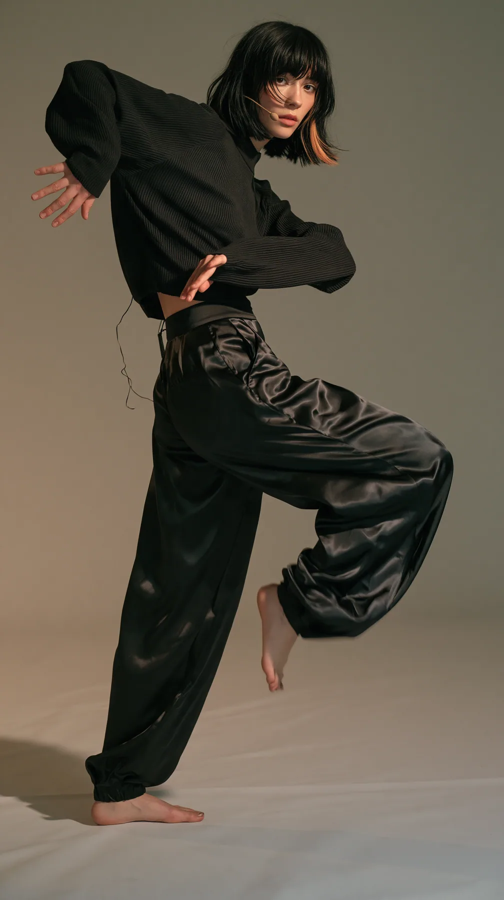 full-length shot, a Caucasian American woman in her late twenties with pale skin and a glossy black blunt bob with heavy straight bangs and one clay-orange streak, a thin headset microphone, in a black ribbed…