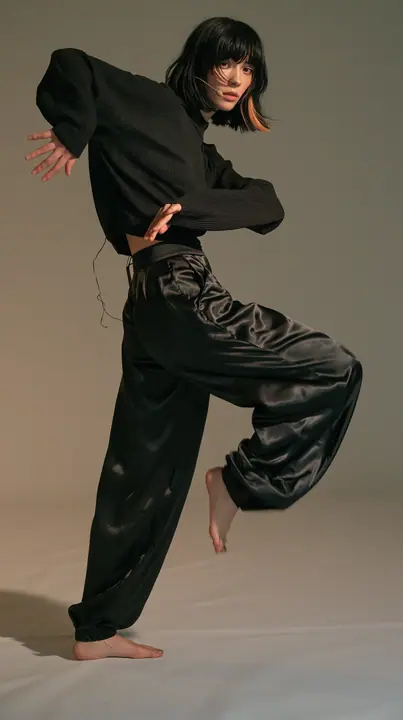 full-length shot, a Caucasian American woman in her late twenties with pale skin and a glossy black blunt bob with heavy straight bangs and one clay-orange streak, a thin headset microphone, in a black ribbed…