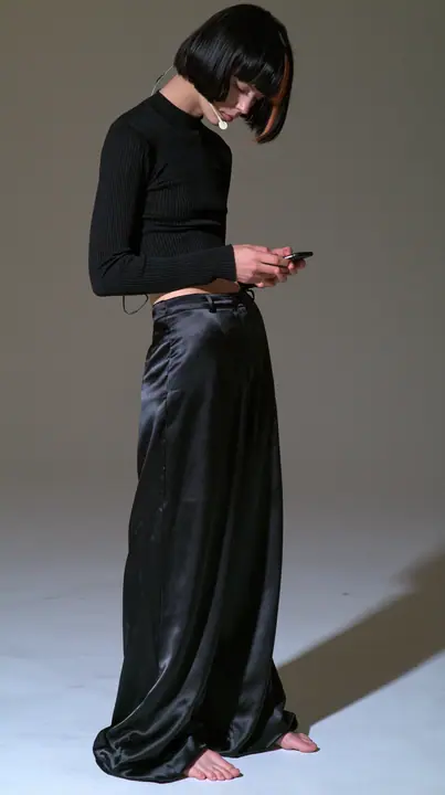 full-length shot, a Caucasian American woman in her late twenties with pale skin and a glossy black blunt bob with heavy straight bangs and one clay-orange streak, a thin headset microphone, in a black ribbed…