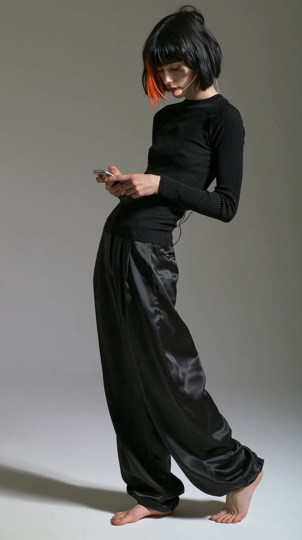 full-length shot, a Caucasian American woman in her late twenties with pale skin and a glossy black blunt bob with heavy straight bangs and one clay-orange streak, a thin headset microphone, in a black ribbed…