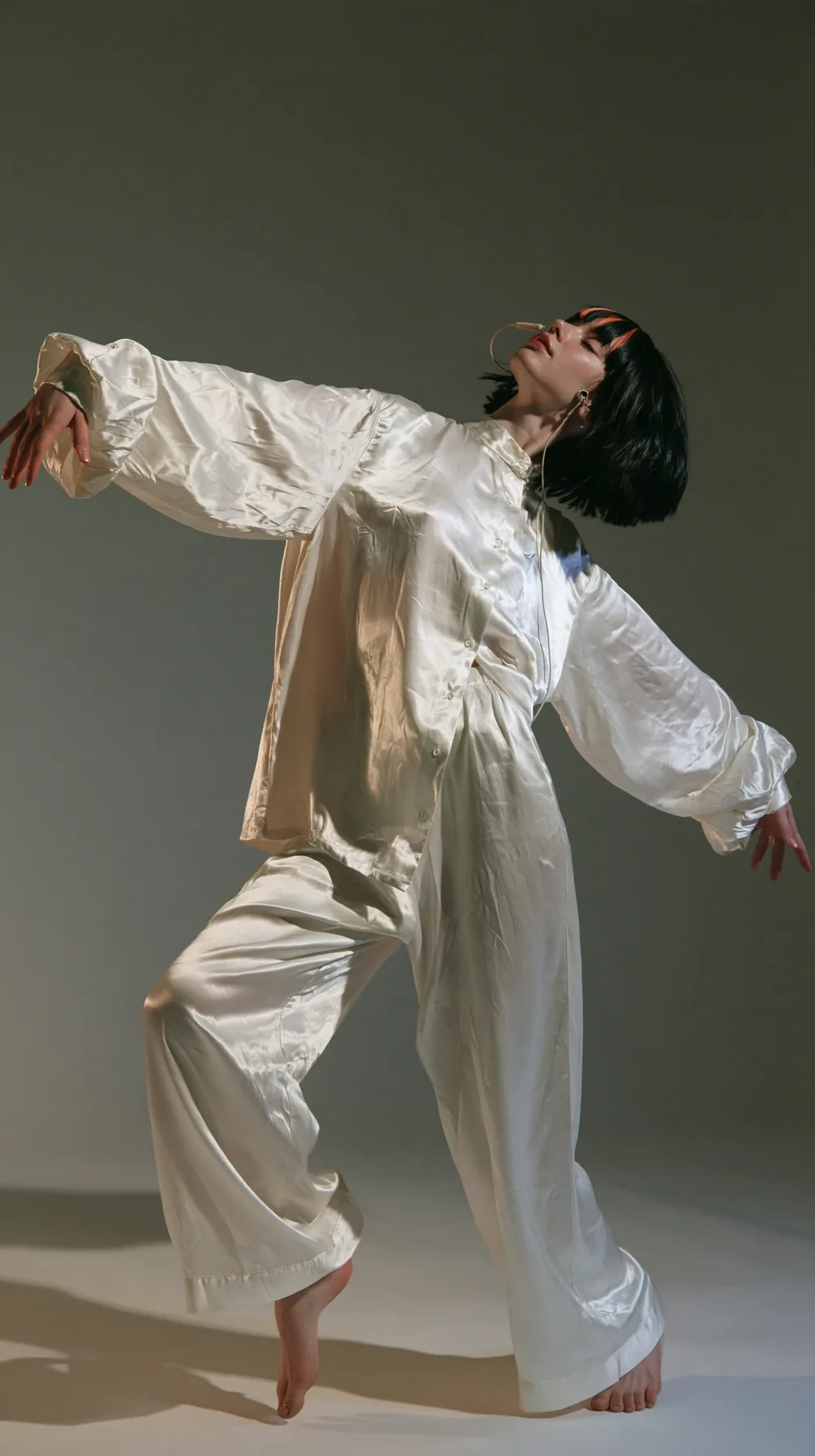 full-length shot, a Caucasian American woman in her late twenties with pale skin and a glossy black blunt bob with heavy straight bangs and one clay-orange streak, a thin headset microphone, in oversized white…